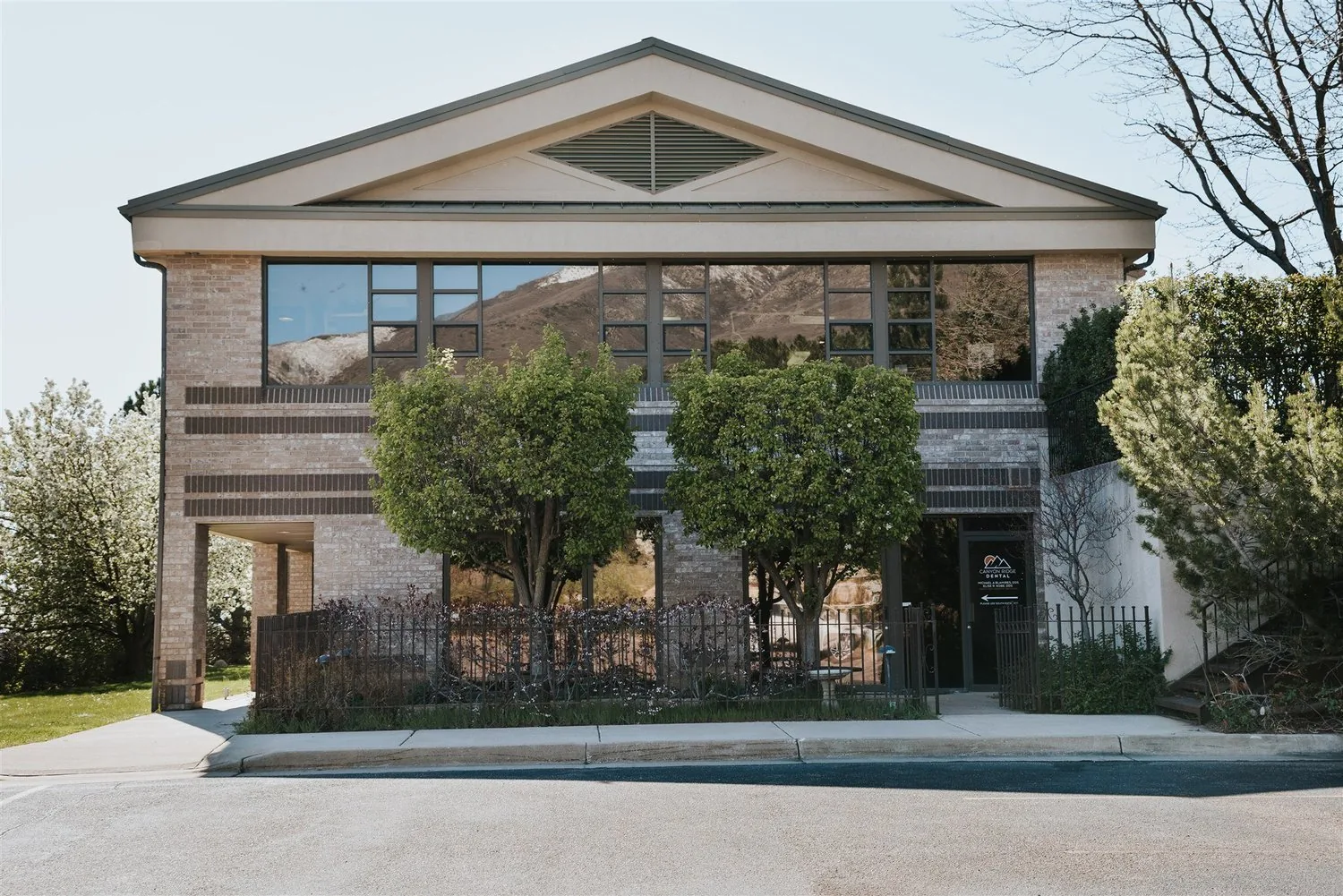 Two-story brick office building with trees out front and large windows reflecting mountains.